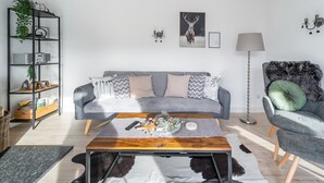 A modern living room with a grey sofa, a wooden coffee table, a black and white cowhide rug, a tall floor lamp, and a side chair with a green cushion.