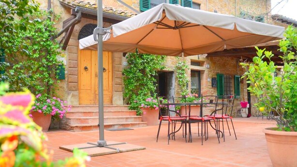 Un patio al aire libre con una mesa y sillas bajo un paraguas grande, rodeado de plantas en macetas y un edificio de piedra con puerta de madera.