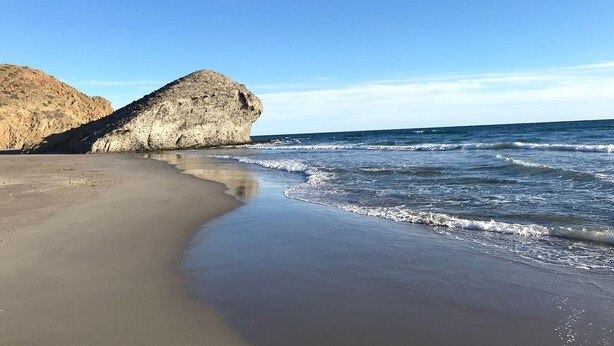 A beach with a large rock formation and calm waves.