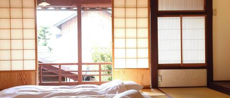 A traditional Japanese room with tatami flooring, a bed with white bedding, sliding doors, and a view of the outside.