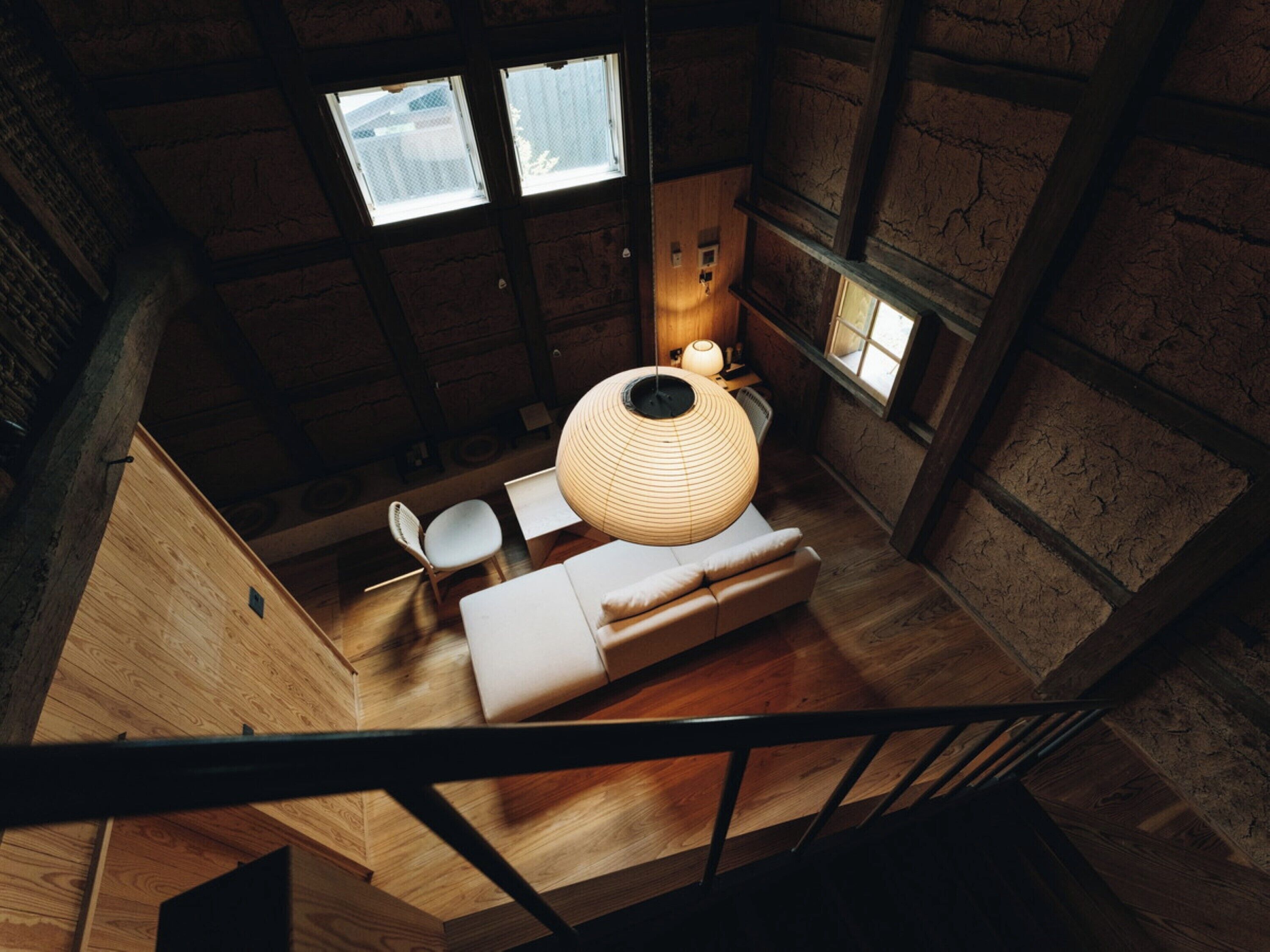 A high-ceiling room with a wooden floor, a round woven light fixture, a sofa, and a chair.