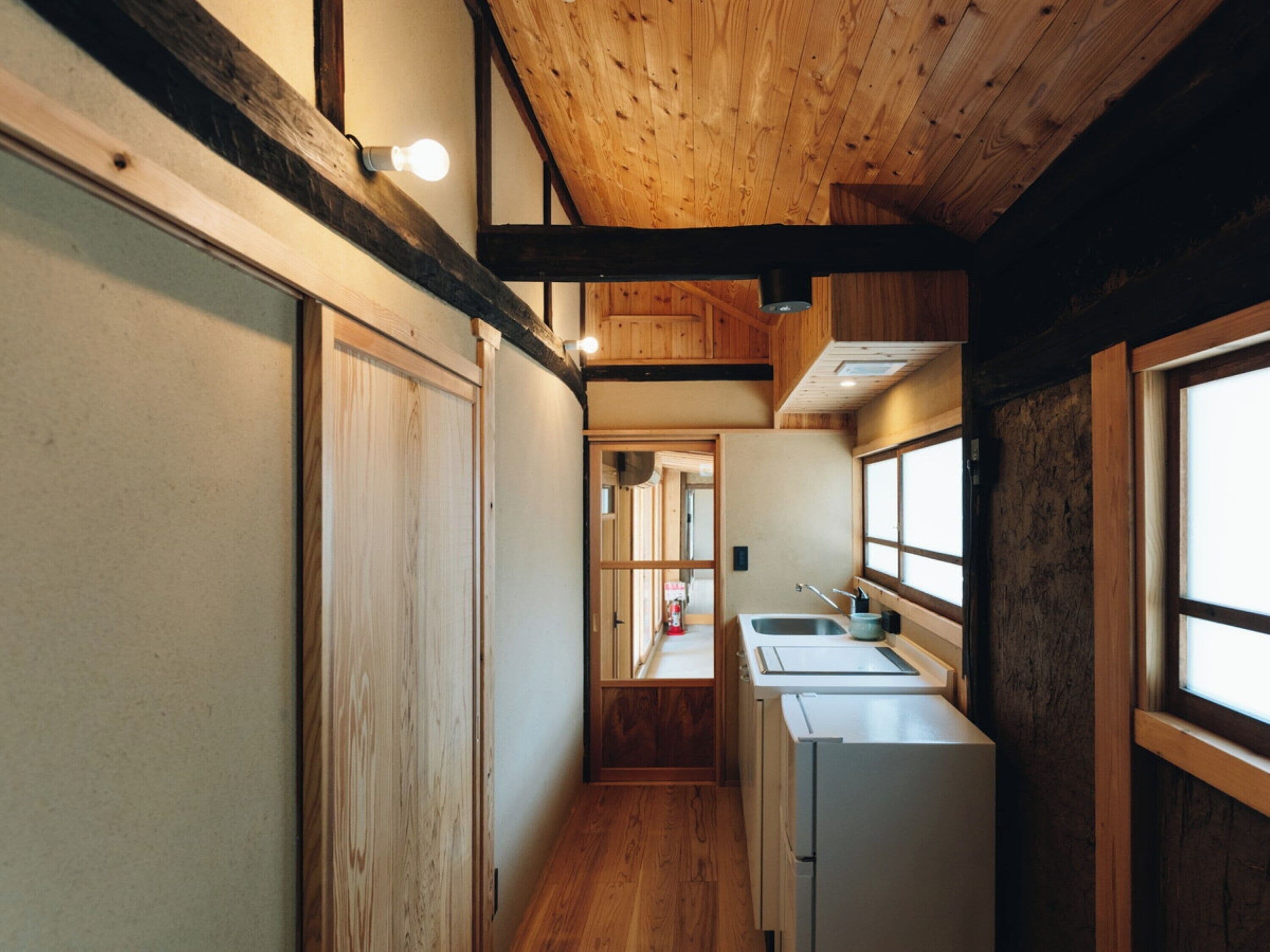 A narrow hallway with wooden walls and a wooden ceiling. There is a small kitchen area with a refrigerator, sink, and cabinets.