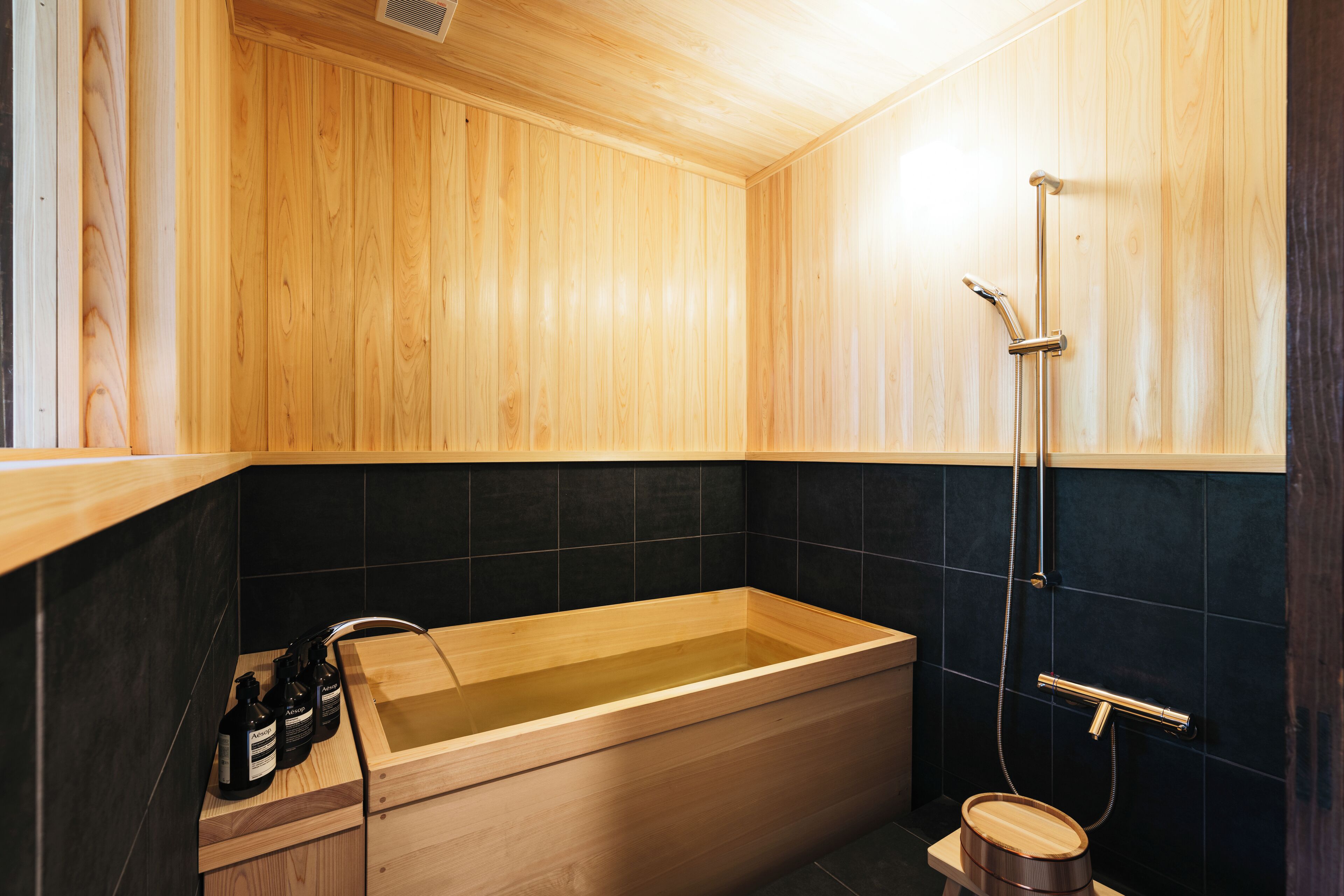 Una bañera de madera con ducha, un banco de madera y baldosas negras.