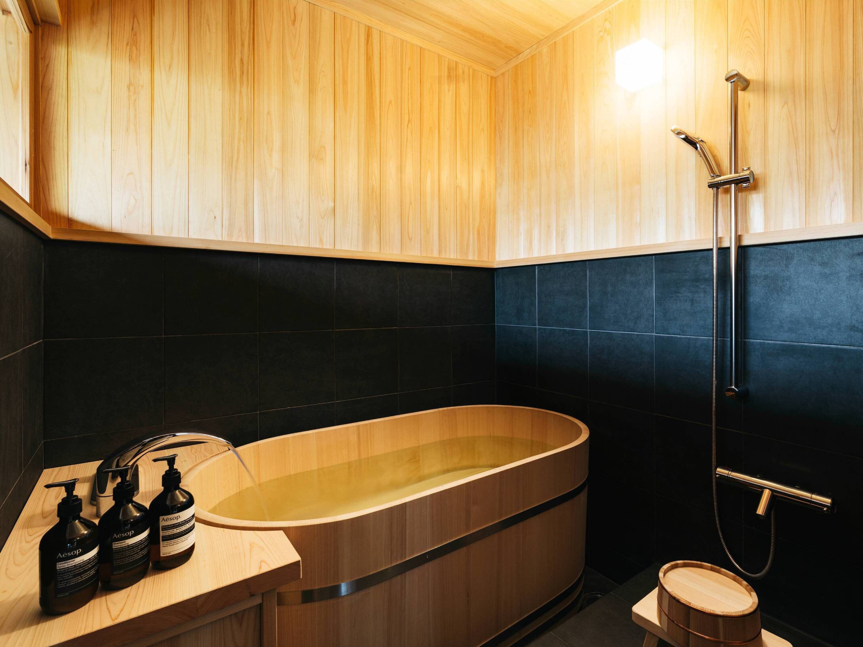 A modern bathroom with a wooden bathtub, black tiled walls, and a wooden vanity with toiletries.