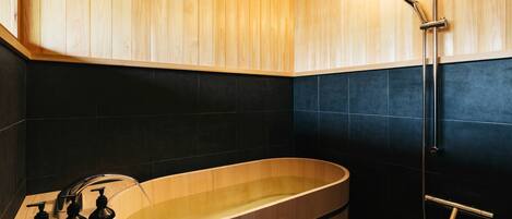 A modern bathroom with a wooden bathtub, black tiled walls, and a wooden vanity with toiletries.