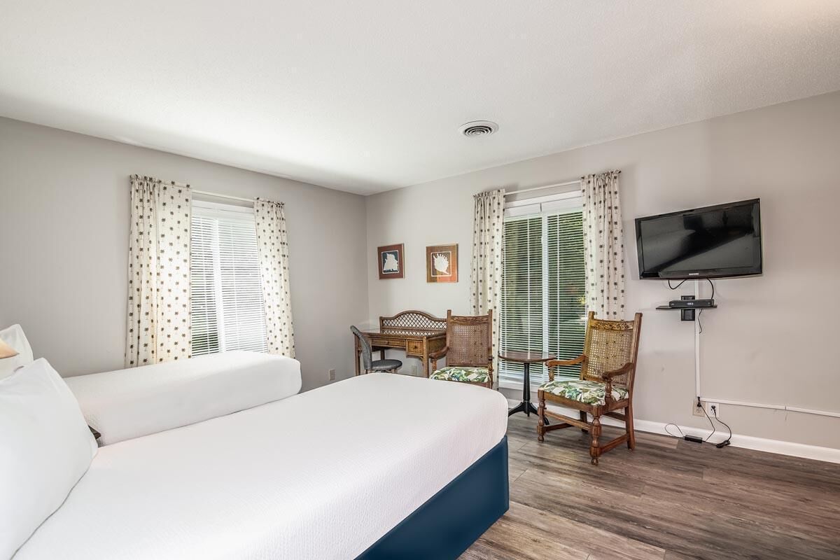 A hotel room with a large bed, two wooden chairs, a television, and two windows with curtains.