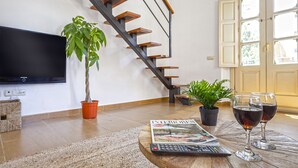 Una sala de estar con una escalera moderna, un televisor de pantalla plana, plantas en macetas, una revista y copas de vino sobre una mesa de madera.