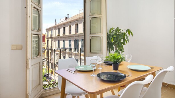 Un comedor con una mesa dispuesta para dos, una planta en maceta y vistas a un edificio a través de ventanas abiertas.