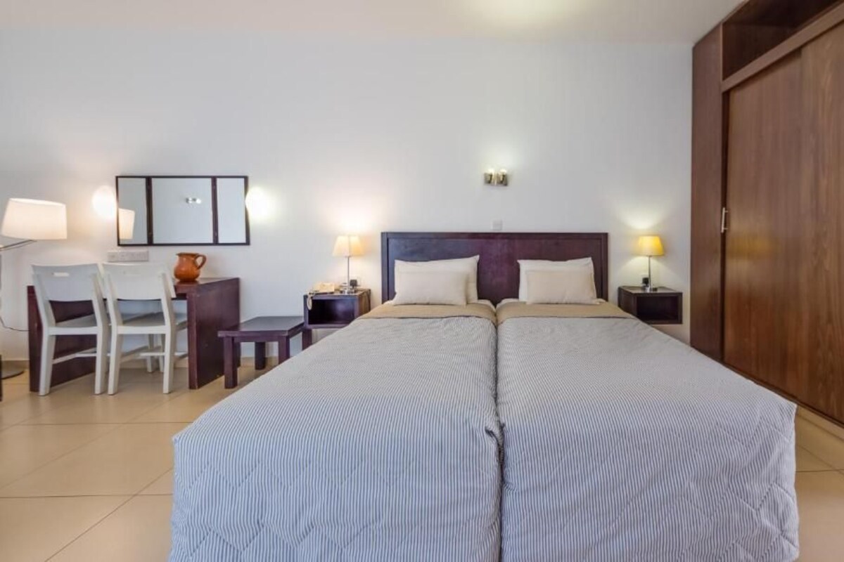Cama doble con cabecera, mesitas de noche con lámparas, escritorio con silla y armario.