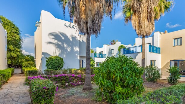 Un'area residenziale moderna con edifici bianchi, balconi blu e vegetazione lussureggiante, tra cui palme e piante fiorite.