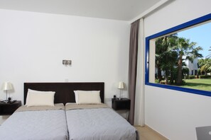 Ein Hotelzimmer mit einem großen Bett, zwei Nachttischen inklusive Lampen, einem Fenster mit Blick auf Palmen und einem blauen Fensterrahmen.