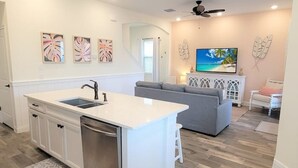 Une cuisine moderne avec un îlot central, un coin repas doté d’un canapé gris et un salon avec un téléviseur et des œuvres d’art décoratives au mur.