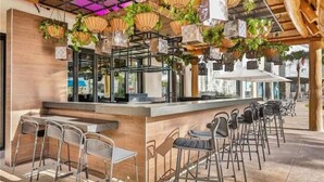 Un espace bar extérieur avec des chaises et des tables en métal, des plantes suspendues et une structure de plafond en bois.