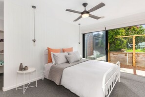 Ein Schlafzimmer mit Bett, Deckenventilator, kleinem Tisch mit Lampe und Blick ins Freie durch eine Schiebetür.