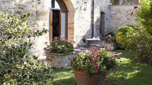 A stone building with an open doorway, a small table with chairs, and potted plants with flowers. - GARDEN HOUSE (San Gimignano)