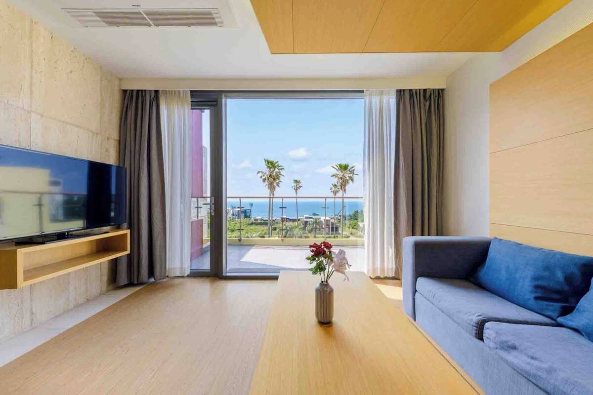 A modern living room with a flat-screen TV, a sofa, a wooden floor, and a view of the ocean through large glass doors.
