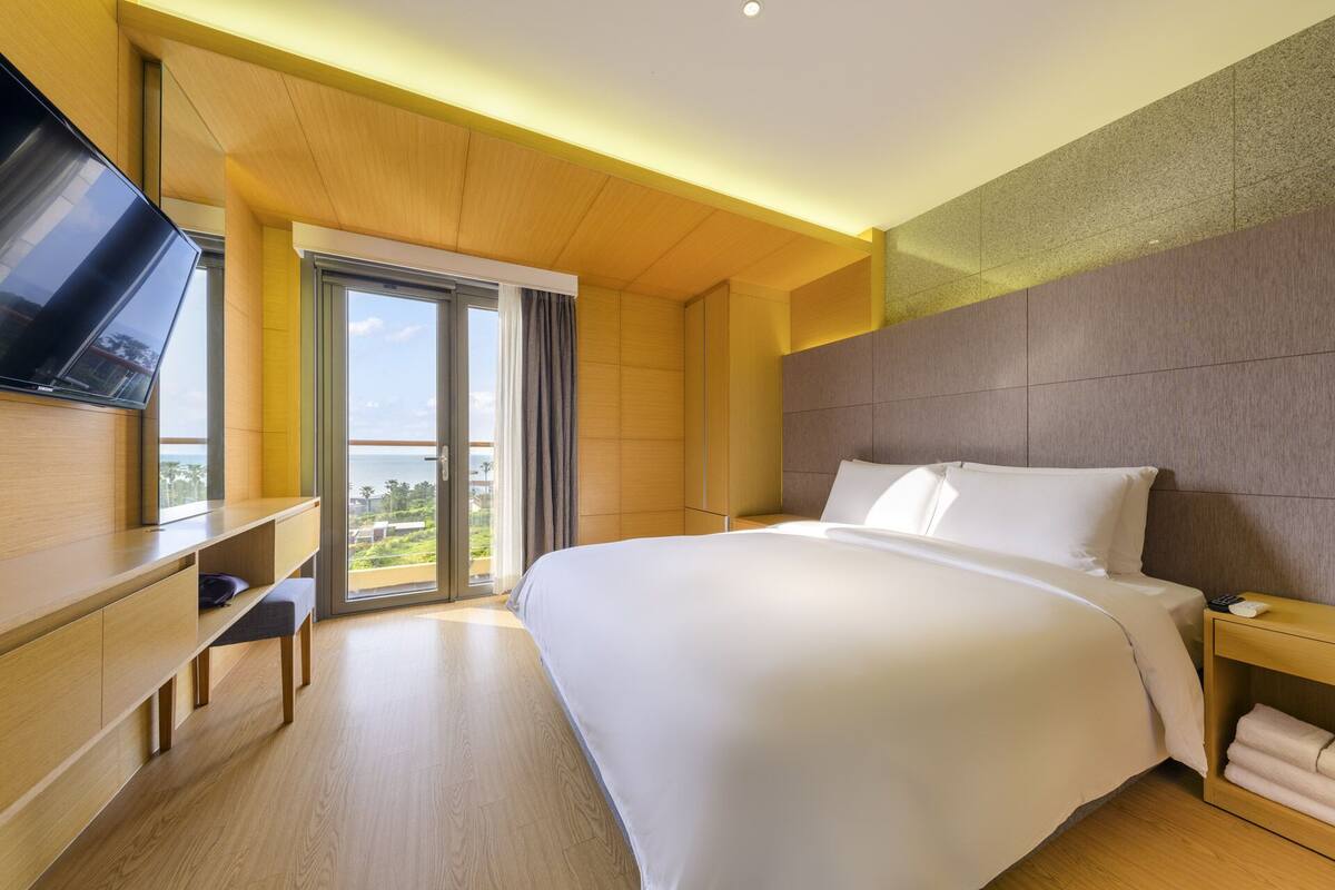 A modern hotel room with a large bed, a desk, a flat-screen TV, and a balcony with a view of the sea.
