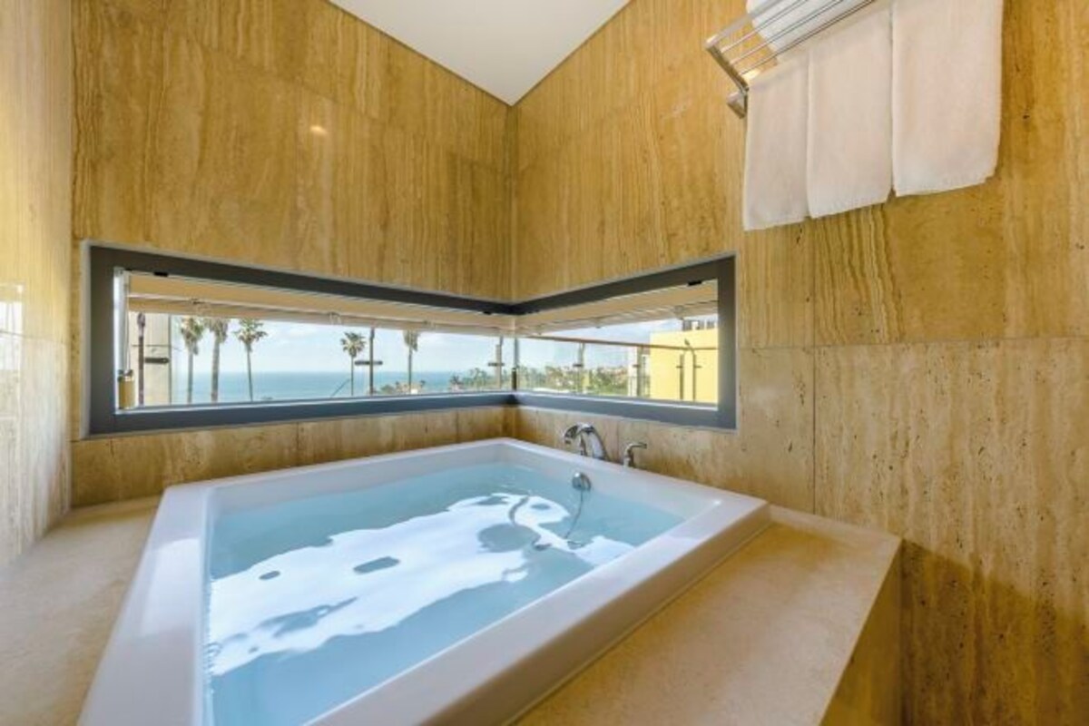 Een moderne badkamer met een groot bad, houten wanden en uitzicht op palmbomen door een raam.