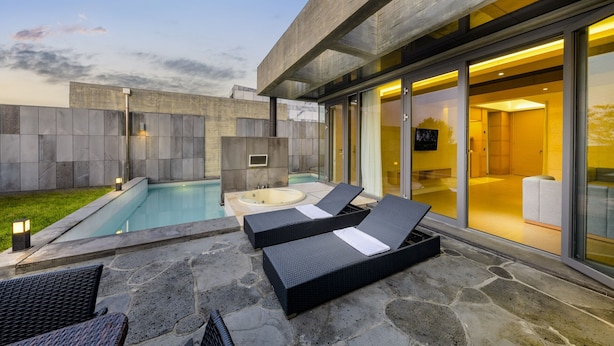 Un área de piscina moderna al aire libre con sillones de descanso y un jacuzzi, rodeada de muros de piedra y puertas de cristal que dan a un espacio interior.