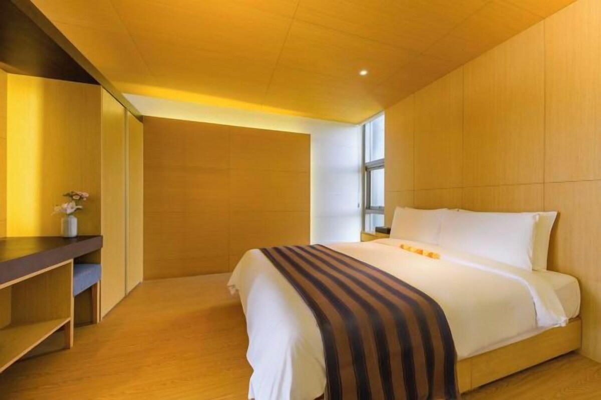 Ein modernes Hotelzimmer mit Holzboden, einem großen Bett mit weißer Bettwäsche, einem Nachttisch mit einer Vase und einem großen Fenster, das natürliches Licht hereinlässt.