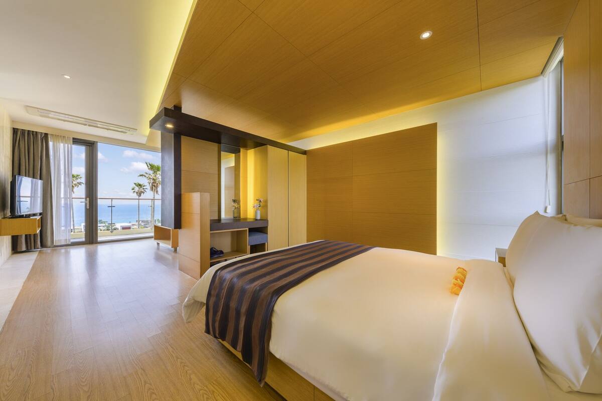 A modern hotel room with a large bed, a TV, a balcony with a view of the ocean, and a wooden ceiling.