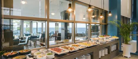 A buffet setup with various food items and a view of the sea through large windows.