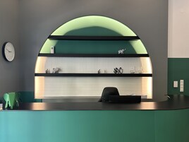 Un mostrador de recepción con una superficie de vidrio curvada, un reloj y una estantería con objetos decorativos.