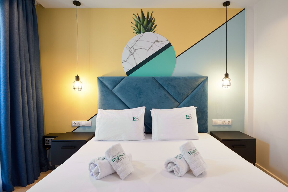 A hotel room with a bed, two pillows with 'ES' logo, a headboard with a pineapple decoration, and two bedside lamps.