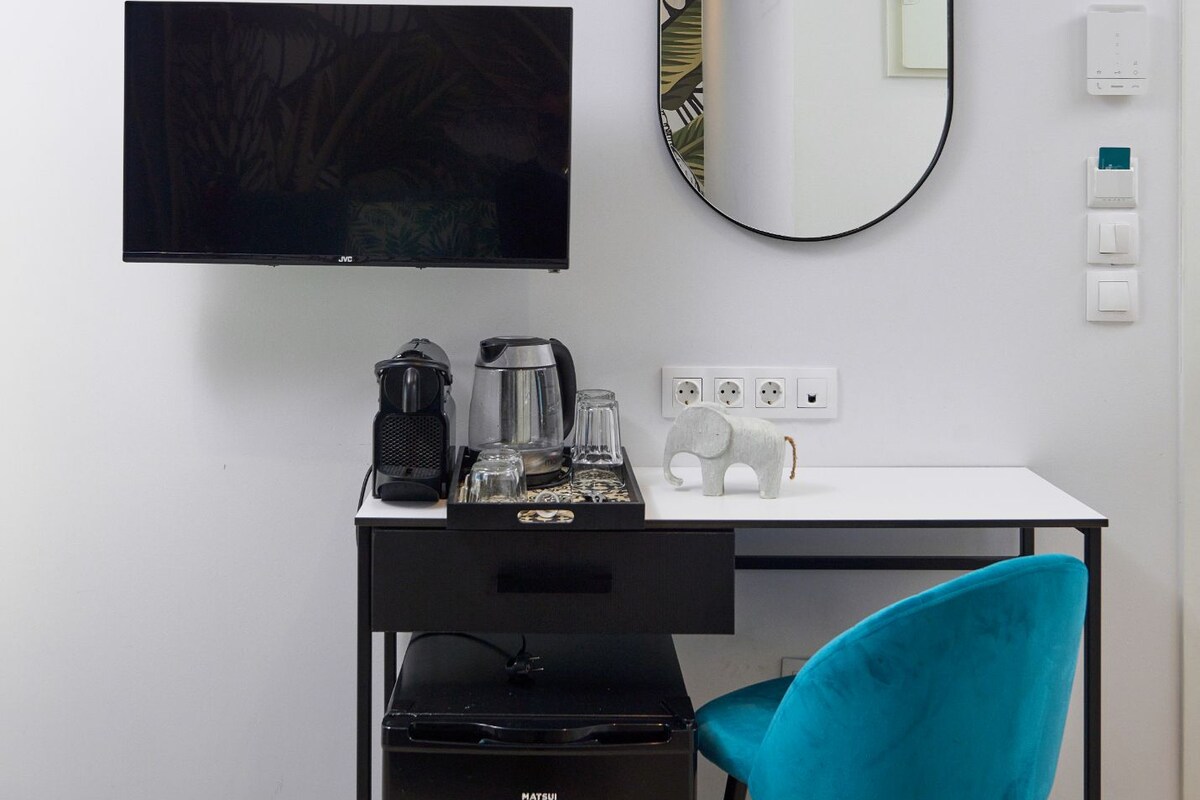 Ein modernes Zimmer mit Fernseher, großem Spiegel, Schreibtisch mit Kaffeemaschine, Laptop und blauem Sessel.