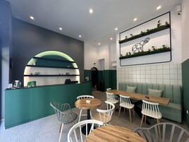 Interior de una cafetería moderna con una paleta de colores verde y blanco, que incluye un mostrador de recepción, un área de estar con mesas y sillas, y un arreglo de plantas en la pared.