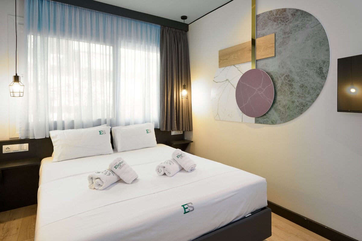 Quarto de hotel com uma cama, duas almofadas com o logotipo 'ES', uma obra de arte na parede e uma mesa de cabeceira com abajur.