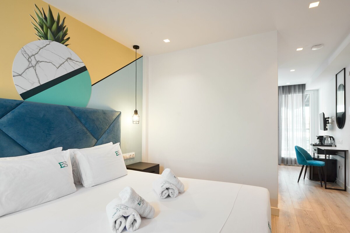 A modern hotel room with a large bed, a desk with a chair, a mirror, and a plant.