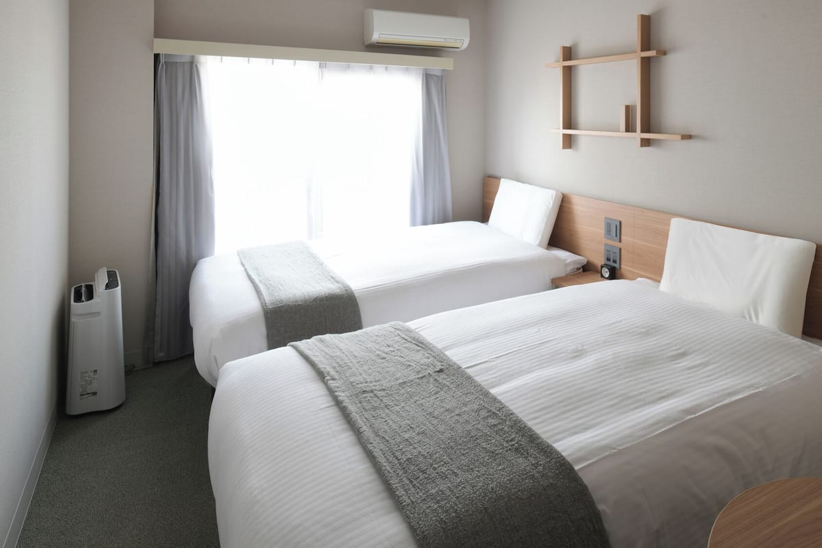 A hotel room with two beds, a bedside table, a wall-mounted shelf, and a window with curtains.