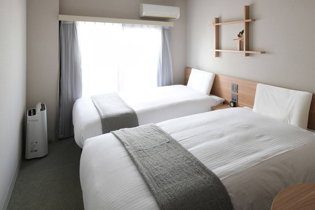 A hotel room with two beds, a wooden headboard, a small wooden shelf, a wall-mounted air conditioner, and a window with curtains.