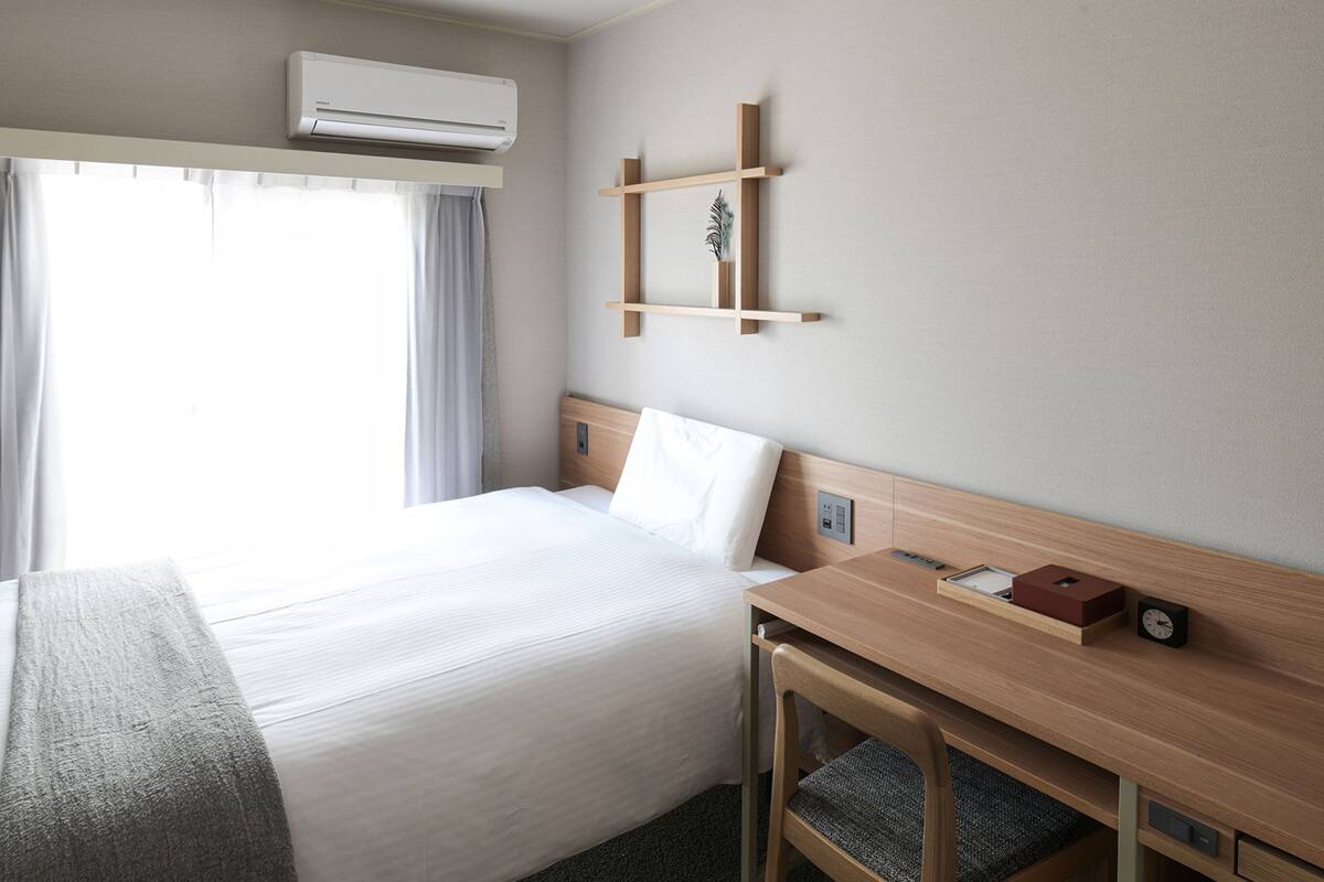 A hotel room with a bed, a desk, a chair, an air conditioning unit, a clock, and a shelf with a plant.