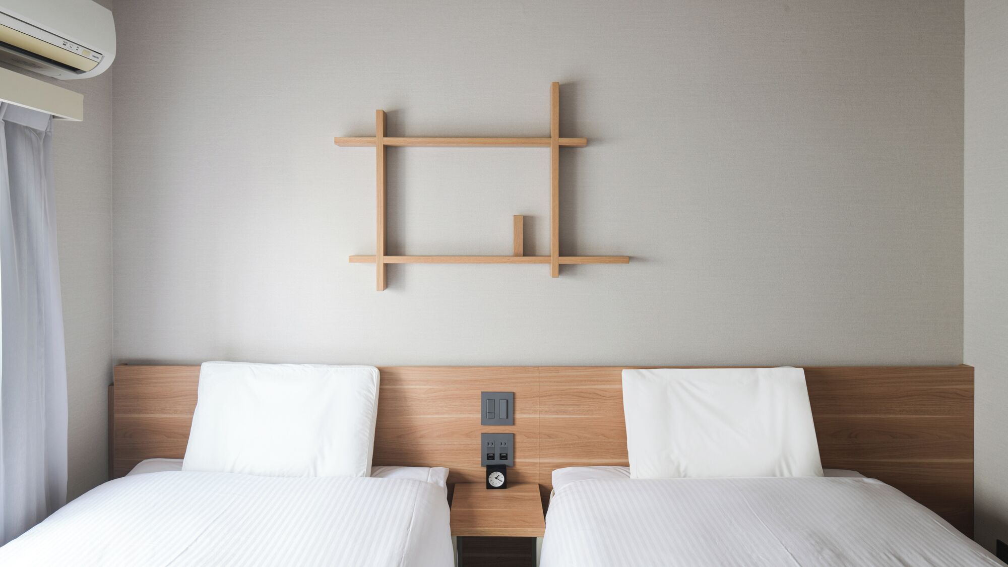 A hotel room with two beds, a wooden headboard, a wall-mounted shelf, and a wall-mounted air conditioning unit.