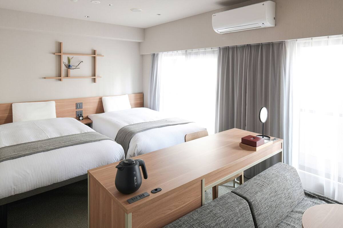A hotel room with two beds, a wooden desk with a kettle, a small table with a mirror, and a wall-mounted air conditioner.