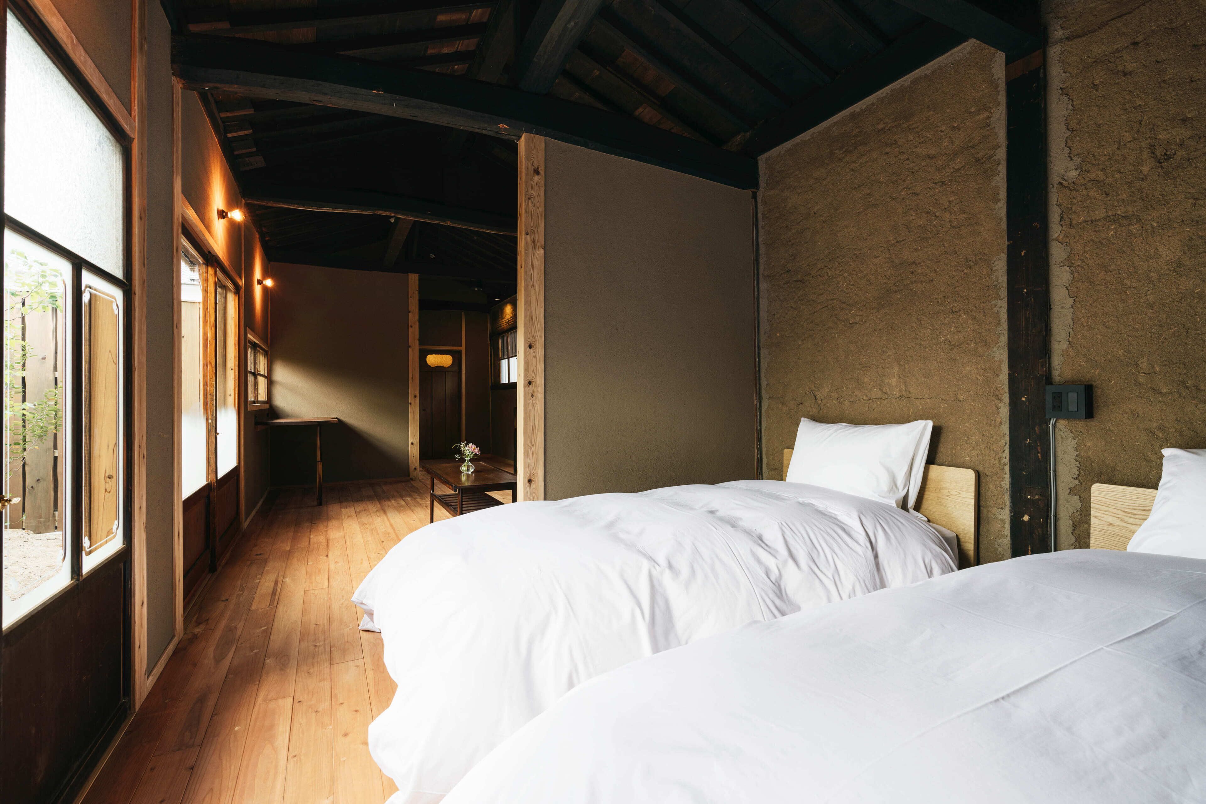 A room with two beds, wooden flooring, a rustic wall, and a small table with a vase.