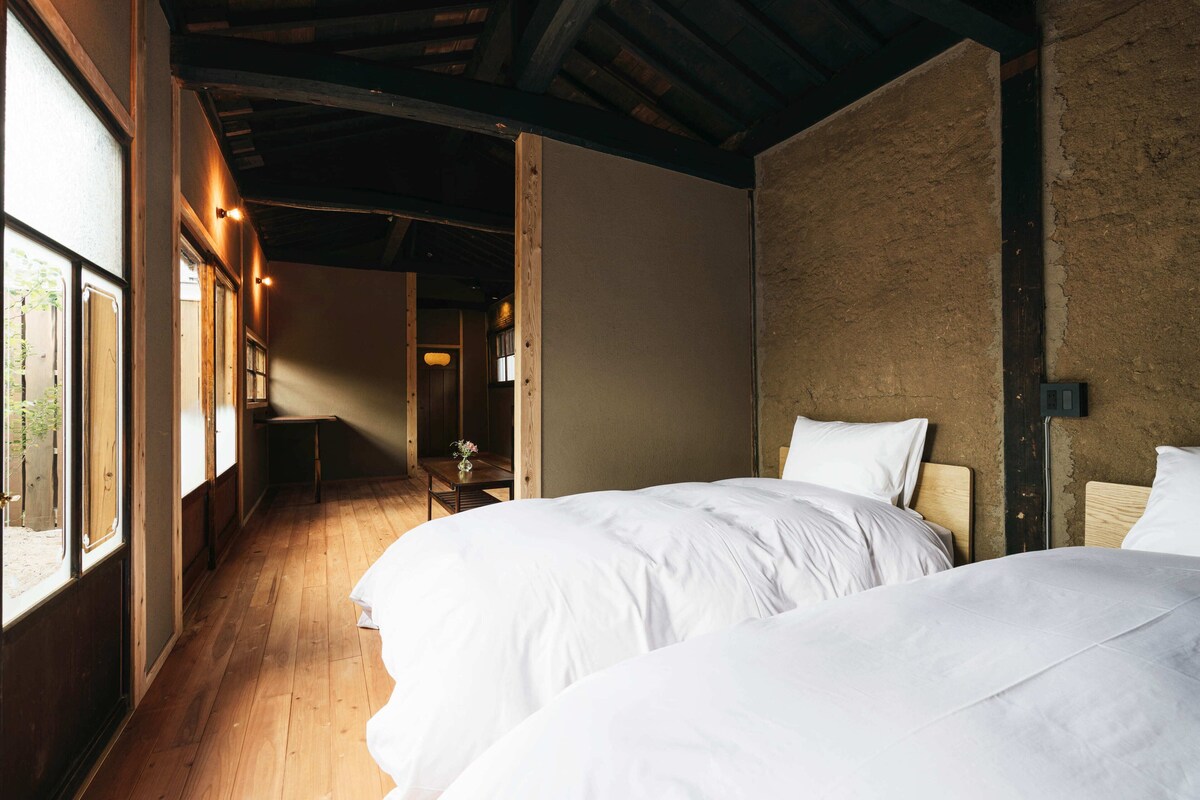 A room with two beds, wooden flooring, a rustic wall, and a small table with a vase.