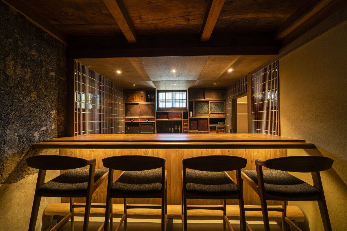 A bar area with wooden chairs and a bar counter, featuring a rustic stone wall and wooden ceiling beams.