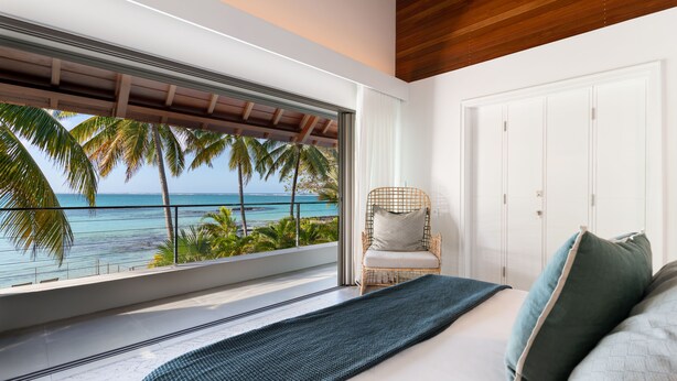 Een slaapkamer met een groot raam met uitzicht op een strand, een bed met kussens, en een rieten stoel.
