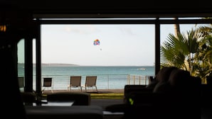 Uitzicht vanuit een kamer met een groot raam op een strand. Drie stoelen staan op het terras en een palmboom aan de rechterkant.