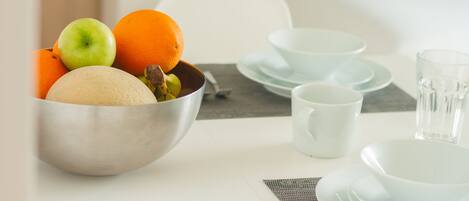 A breakfast setting with a bowl of fruit, including an apple, orange, and melon, on a table with white plates and a cup.