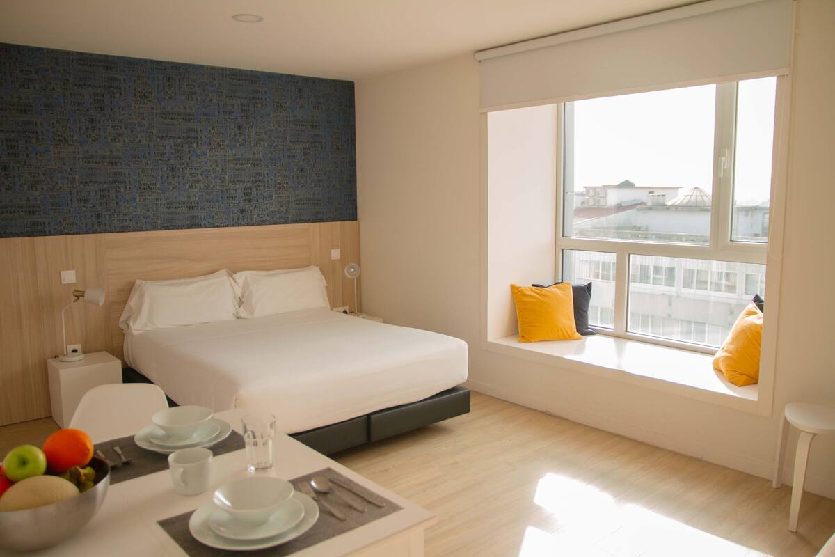 A modern hotel room with a bed, a dining area with a table set for two, and a large window with a city view.