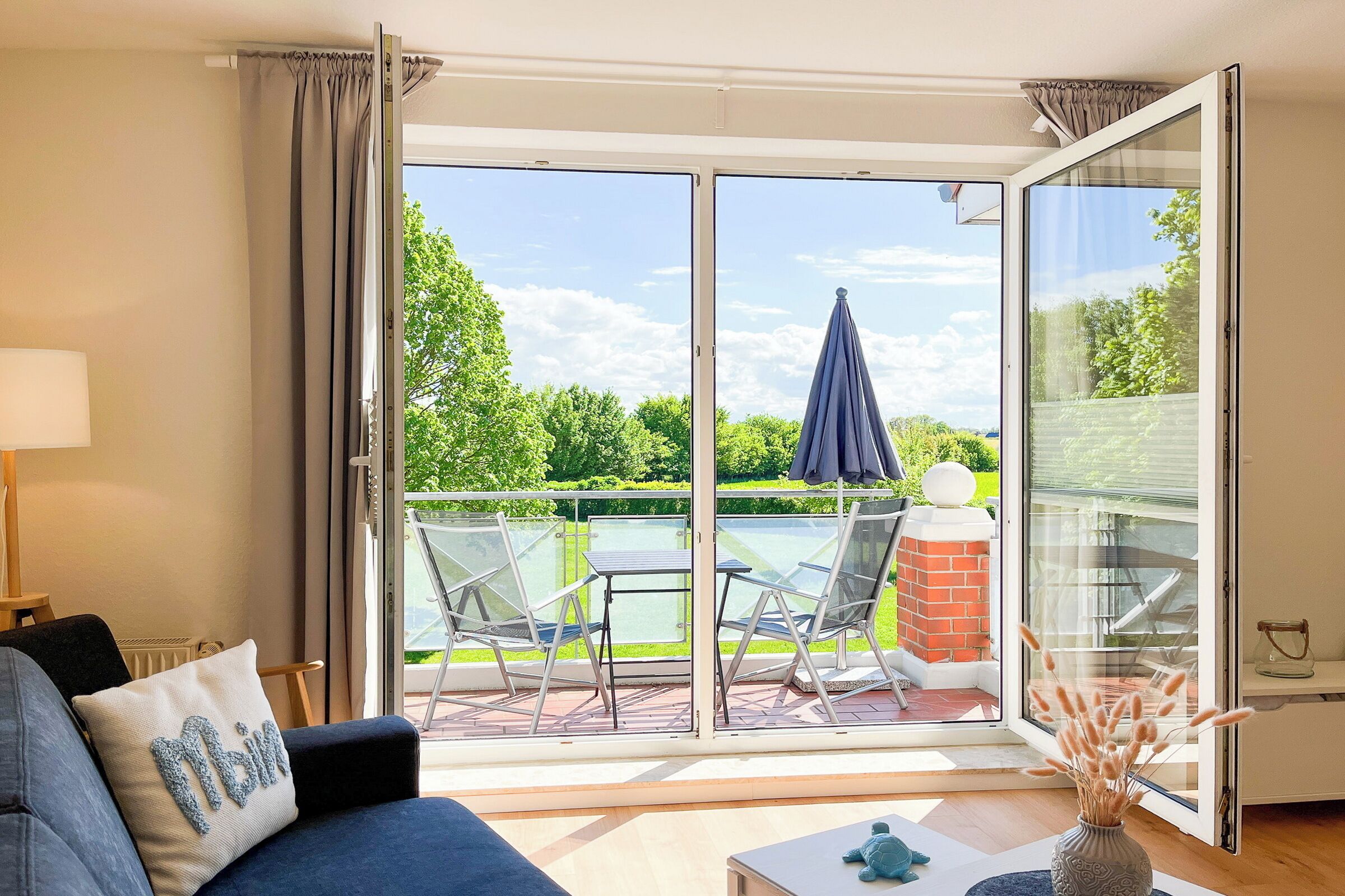 A living room with a sofa, a table, and a view of an outdoor patio with a glass door, a chair, and a table.