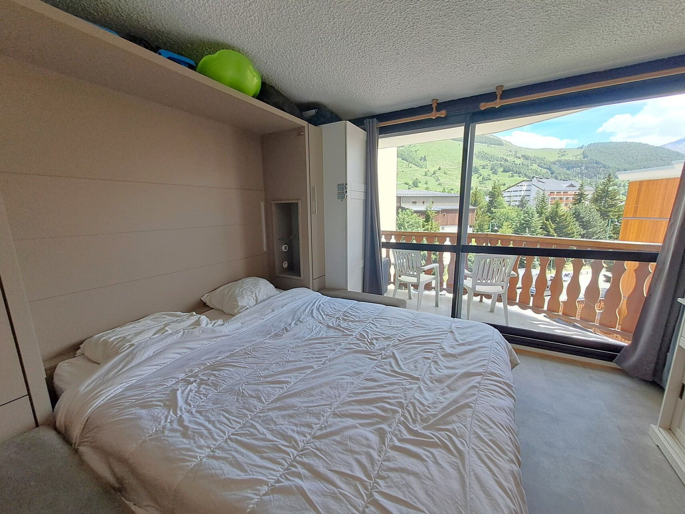 A bedroom with a bed, a window, and a view of a balcony with chairs.