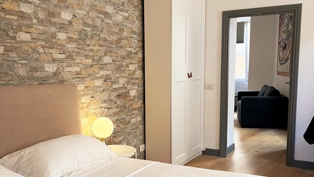 A hotel room with a stone wall, a bed with white linens, a wardrobe, and a hallway leading to another room. - APARTMENT 4 PANTHEON (Roma)