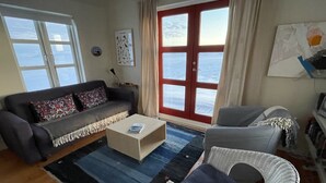 Sala de estar con sofá, una mesa de centro, una silla de mimbre y vista al mar a través de una puerta roja.