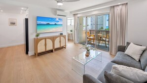 Ein modernes Wohnzimmer mit Flachbildfernseher, einem Glastisch, einem grauen Sofa und einem Balkon mit Meerblick.