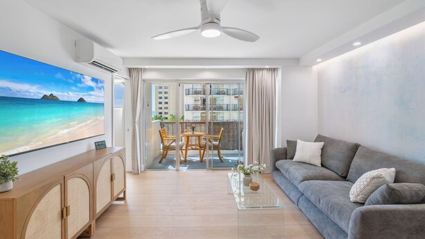 Ein modernes Wohnzimmer mit einem grauen Sofa, einem Glastisch und einem großen Flachbildfernseher, der eine Strandansicht zeigt.